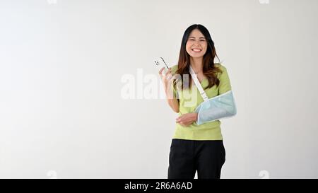 Una giovane asiatica allegra e sorridente con un braccio rotto, indossando un'imbracatura per sostenere il braccio ferito e tenendo il suo smartphone. isolato su un Foto Stock