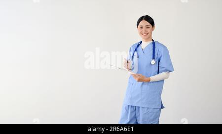 Un attraente e affascinante giovane medico asiatico o operatrice medica in scrub tenendo una clipboard medica, mentre in piedi contro un bianco isolato Foto Stock