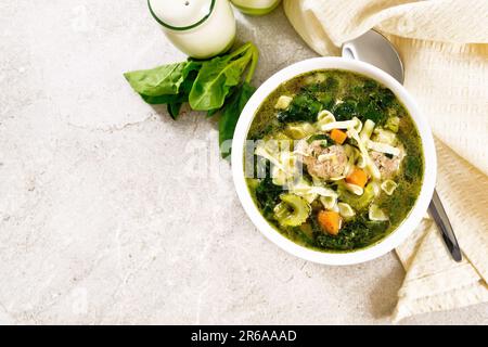 Ministra maritata con polpette, spinaci, carote, sedano e pasta fine in una ciotola, tovagliolo e cucchiaio su fondo di pietra ta Foto Stock