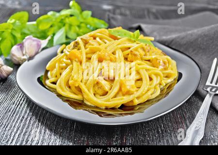 Spaghetti con salsa di zucca, pancetta, cipolla e aglio in un piatto, asciugamano da cucina, basilico e forchetta su fondo di legno Foto Stock