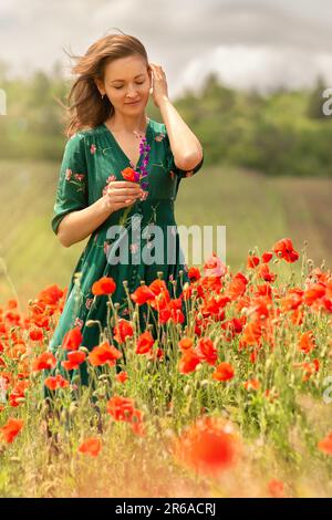 una giovane donna bianca in un campo di papavero, con una mano raddrizza i capelli, nell'altra tiene un fiore rosso con un sorriso, un'atmosfera di gioia, amore Foto Stock