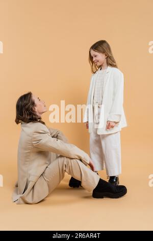 madre bambino legame concetto, donna elegante in tuta seduta e guardando felice preseen figlia su sfondo beige, madre aziendale, donna d'affari, m Foto Stock