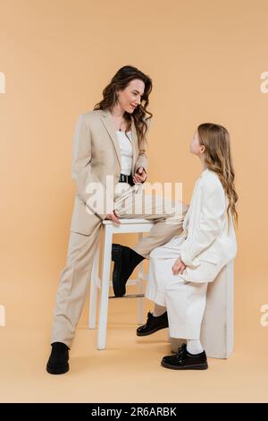 mamma e figlia corporativa in tute, donna seduta su seggiolone e guardando la ragazza su sfondo beige, abiti alla moda, abbigliamento formale, corporat Foto Stock