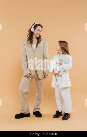 madre e figlia in tute, donna in cuffie wireless e ragazza con notebook su sfondo beige, abiti alla moda, abbigliamento formale, mamma aziendale Foto Stock