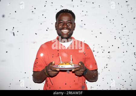 Minimal vita up ritratto di uomo nero sorridente che tiene torta di compleanno con confetti doccia alla festa, girato con il flash Foto Stock