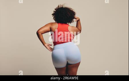 Vista posteriore di una donna fiduciosa in piedi con sicurezza in uno studio, vestita di abbigliamento sportivo e che mostra il suo corpo fisico in forma. Sport giovane donna embracin Foto Stock