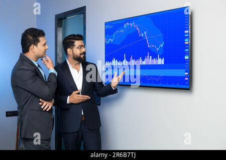 Uomini d'affari indiani colleghi che commerciano e discutono gli stock in linea in ufficio. Broker che guardano i grafici degli indici sul grande schermo del computer. Foto Stock