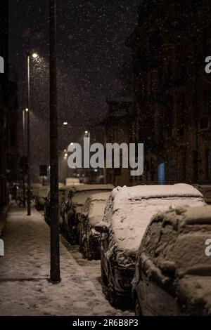 Un paesaggio urbano innevato è mostrato con le automobili parcheggiate su un marciapiede e le persone che camminano sullo sfondo Foto Stock