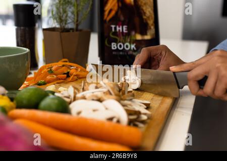 Parte centrale della donna biraciale preparare il cibo in cucina tritare le verdure e usare la compressa Foto Stock