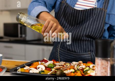 Sezione centrale della donna biraciale in grembiule preparazione del cibo in cucina versando olio sulle verdure Foto Stock