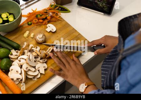 Parte centrale della donna biraciale preparare il cibo in cucina tritare le verdure e usare la compressa Foto Stock