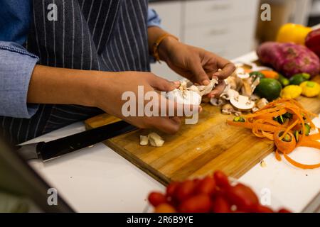 Parte centrale della donna biraciale in grembiule preparare il cibo in cucina sbucciare l'aglio Foto Stock