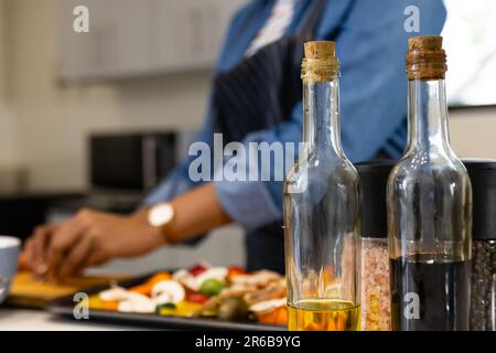 Bottiglie di olio e aceto e la parte centrale della donna biraciale in grembiule preparazione del cibo in cucina Foto Stock