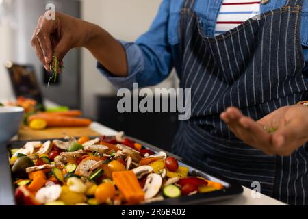 Sezione centrale della donna biraciale in grembiule preparazione del cibo in cucina condimenti vegetali Foto Stock