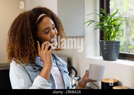 Felice donna biraciale che ha caffè e parlare sullo smartphone in cucina a casa Foto Stock