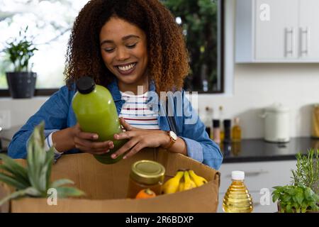 Felice donna biraciale disimballaggio negozio di alimentari da scatola riciclata in cucina Foto Stock