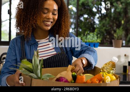 Felice donna biraciale disimballaggio negozio di alimentari da scatola riciclata in cucina Foto Stock