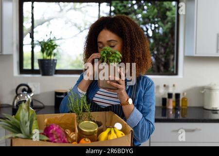 Felice donna biraciale disimballaggio negozio di alimentari dalla scatola in cucina e odori erbe Foto Stock