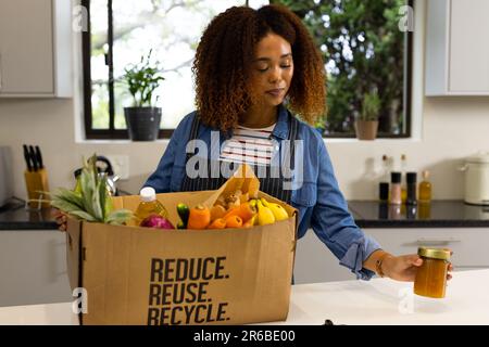 Felice donna biraciale disimballaggio negozio di alimentari da scatola riciclata in cucina Foto Stock