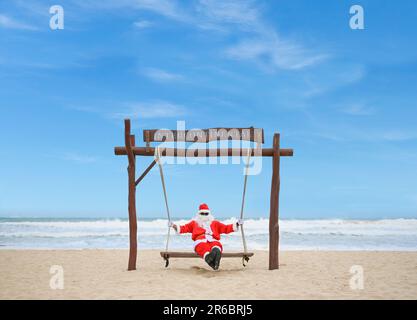 Un Babbo Natale jolly godendosi una giornata alla spiaggia mentre si siede in un altalena. Foto Stock