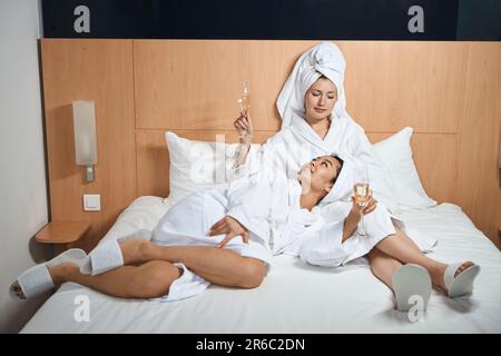 Due amiche felici bevono champagne in accappatoi bianchi in hotel Foto Stock