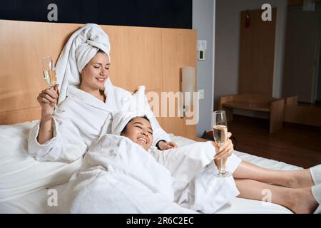 Due donne felici bevono champagne in accappatoi bianchi sul letto Foto Stock