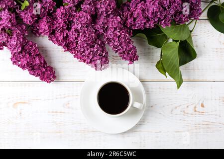 Tazza di caffè nero fresco e ramo di lilla viola fiorente con foglie verdi su sfondo di legno bianco. Pausa caffè. Fiori primaverili. Giacitura piatta, da a. Foto Stock