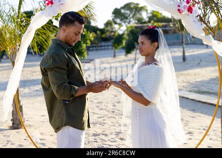 Vista laterale del giovane sposo caucasico che indossa l'anello di nozze per sposarsi durante la cerimonia nuziale in spiaggia Foto Stock