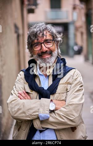 Verticale uomo con capelli grigi incrociati braccia sorridente felice guardando la macchina fotografica in piedi città strada. Foto Stock