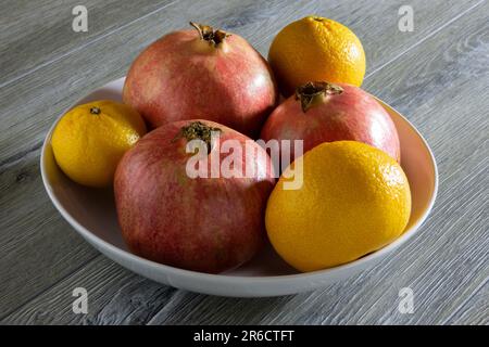 Tre di melograni freschi maturi e tre arance in un piatto su un tavolo di legno grigio. Primo piano Foto Stock