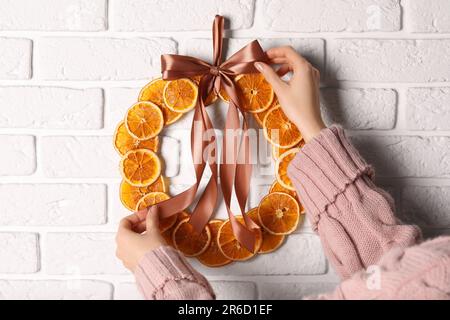 Donna legatura nastro su corona decorativa con arance a secco vicino muro di mattoni bianchi, primo piano Foto Stock
