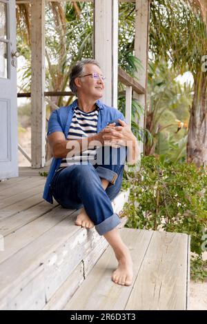 Sorridente uomo caucasico vestito di casuals contemplare mentre seduto su gradini fuori cottage Foto Stock