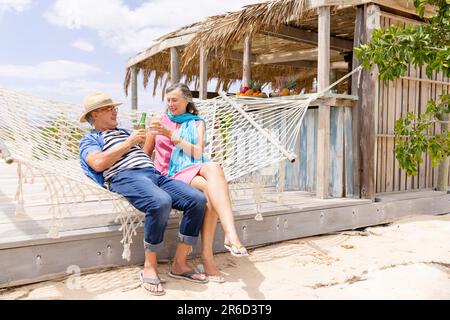 Coppia caucasica anziana che brinda drink e si rilassa sull'amaca in spiaggia Foto Stock