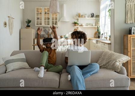 Moderna famiglia afro-americana, mamma e figlio si siedono sul divano con dispositivi elettronici. Tecnologia e bambini Foto Stock