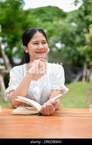 Un ritratto di una bella e sognante giovane donna asiatica in abiti casual sta leggendo un libro a un tavolo all'aperto, ama trascorrere il suo tempo libero al c Foto Stock