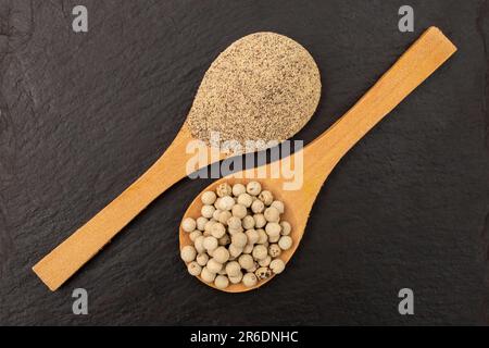 Semi di pepe bianco vs polvere e cucchiai di legno, su mattonelle di pietra nera, piatto si dispongono da vicino Foto Stock