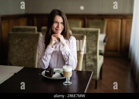 Giovane bella donna brunette sedersi nel bar ristorante caffetteria al coperto e scricchiola il suo volto. Foto Stock