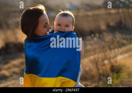 La donna abbraccia il suo piccolo figlio avvolto in giallo e bandiera blu dell'Ucraina all'aperto. Foto Stock