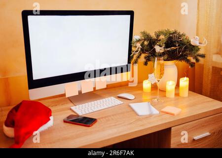 Decorazioni natalizie presso un accogliente posto di lavoro e cappello di santa. Computer con schermo mock-up vuoto, telefono e notebook. Vaso con ramo di albero di Natale, Foto Stock