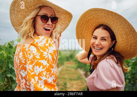 Due giovani donne belle e felici in abiti eleganti e ampi cappelli di paglia brimmed sorridono e guardano la macchina fotografica. Amici donne attrattivi a piedi t Foto Stock