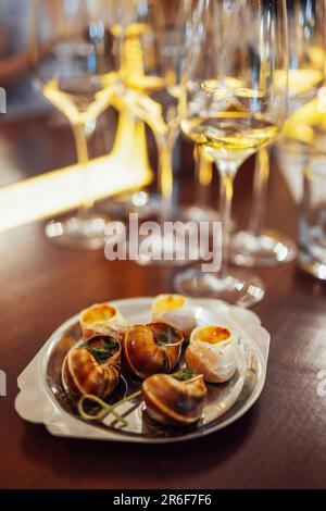 Una squisita delicatezza di lumache su un vassoio di metallo Escargot con erbe su un piatto su un tavolo di legno. Deliziosa lumaca bourguignonne. Bicchieri da vino su ba Foto Stock