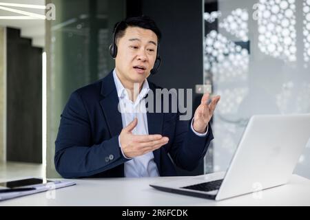 Ritratto di giovane consulente asiatico maschile in servizio di supporto. Seduti in ufficio al tavolo, indossando cuffie e un microfono, parlando con i clienti online da un computer portatile Foto Stock