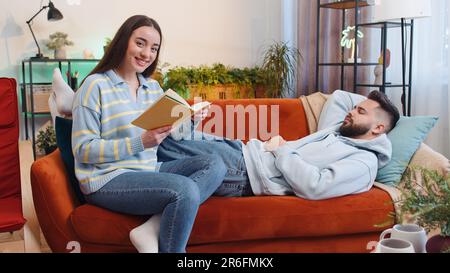 Donna che legge la storia dal libro mentre dispiaciuta l'uomo del ragazzo stanco che si addormenta e russare. Giovane coppia di famiglia a casa. Marito moglie insieme sul divano in soggiorno. Attività ricreative hobby Foto Stock