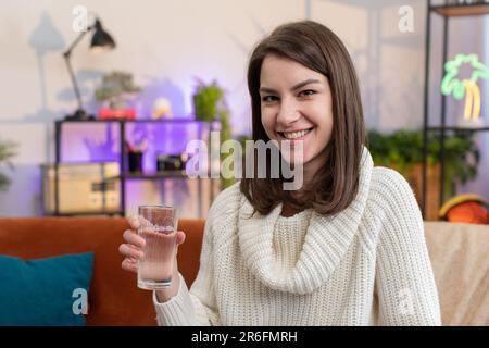 Giovane donna assetata che tiene il vetro di acqua naturale fare sorsi bere ancora acqua impedendo disidratazione si siede nel salotto di casa. Ragazza con buone abitudini di vita, sano dimagrimento, concetto di perdita di peso Foto Stock