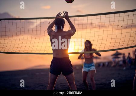 Una giovane coppia gioca a pallavolo sulla spiaggia mentre il sole tramonta in una bella giornata estiva. Sport, mare, spiaggia Foto Stock