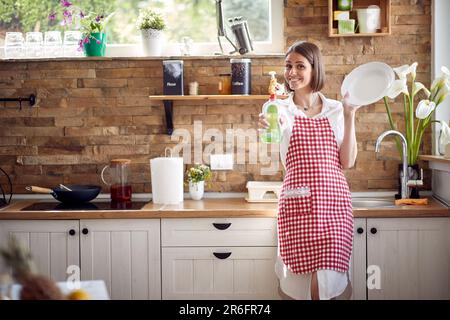 Attraente giovane casalinga che indossa grembiule a scacchi in piedi in cucina, tenendo un piatto pulito e dimostrando il detergente preferito per piatti e. Foto Stock