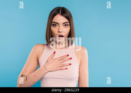 donna scioccata con mano sul petto, modello elegante con tatuaggi e piercing naso guardando la macchina fotografica su sfondo blu, emotivo, bocca aperta, generati Foto Stock