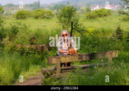 Donna con il suo dachshund marrone a pelo corto in piedi su una panca di legno, guardando la macchina fotografica, la vegetazione selvaggia e gli alberi verdi frondosi su sfondo appannato, Foto Stock