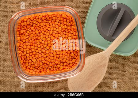 Lenticchie rosse in contenitore di plastica con cucchiaio di legno su tela di iuta, macro, vista dall'alto. Foto Stock