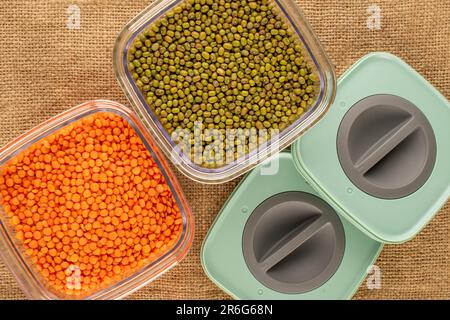 Lenticchie rosse e fagiolini in contenitori di plastica su tela di iuta, macro, vista dall'alto. Foto Stock